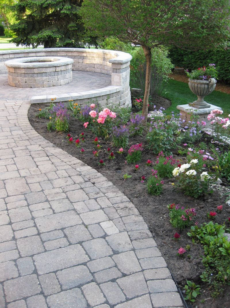 A stone paver walkway curves past a garden bed with colorful flowers toward a circular stone fire pit and seating area.