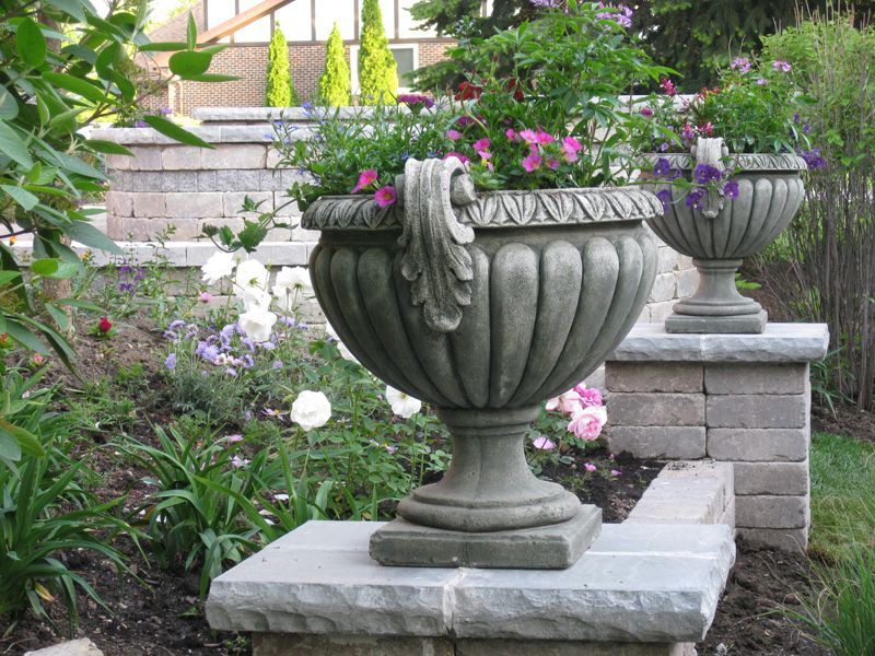 Two stone urn planters filled with colorful flowers sit atop stone pillars in a garden.