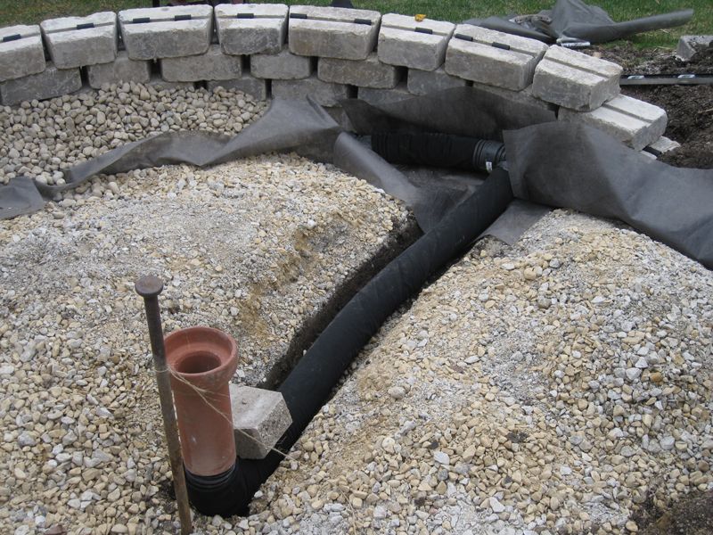 A fire pit under construction featuring a concrete block ring, gravel base, black drainage pipe, and a central vent pipe.