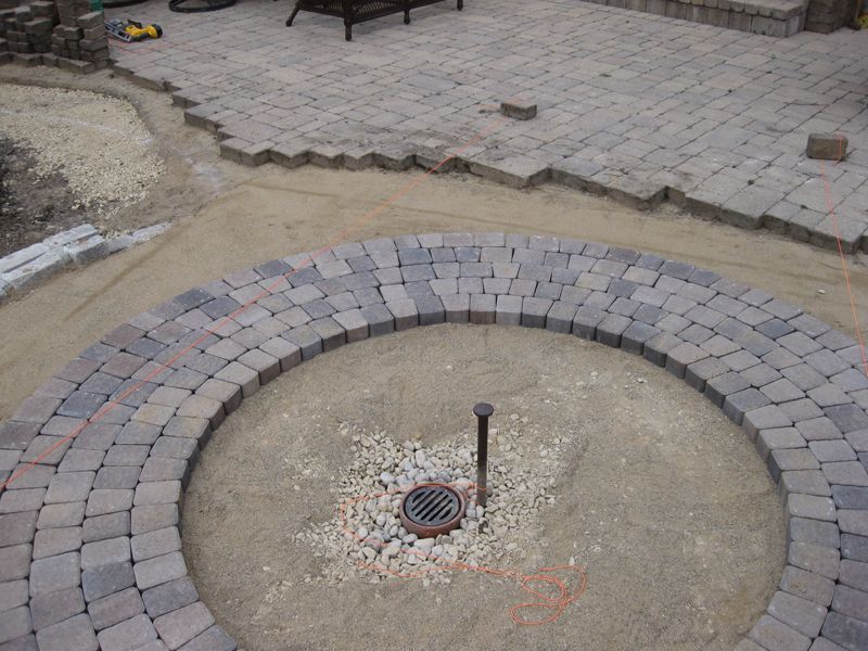 A circular stone border being built on a sand base, featuring a central drainage grate and a metal post.