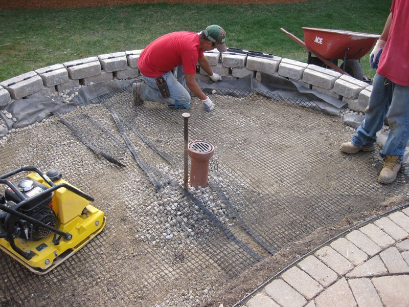 Workers construct a patio base, layering gravel, mesh, and landscape fabric inside a curved block retaining wall.