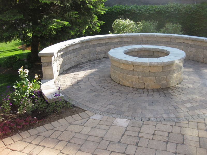 A circular stone fire pit sits on a patterned paver patio, framed by a curved stone retaining wall and lush landscaping.