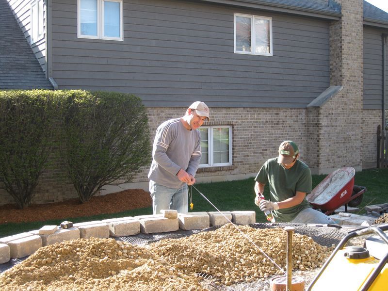 Two workers laying concrete retaining wall blocks on a prepared gravel base in a residential yard.