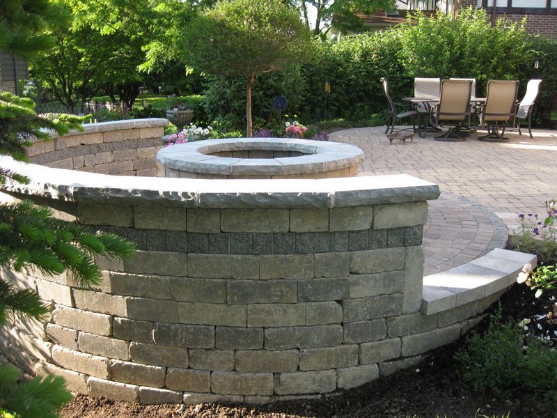 A curved stone seating wall surrounds a stone fire pit on a patio with a dining set in the background.