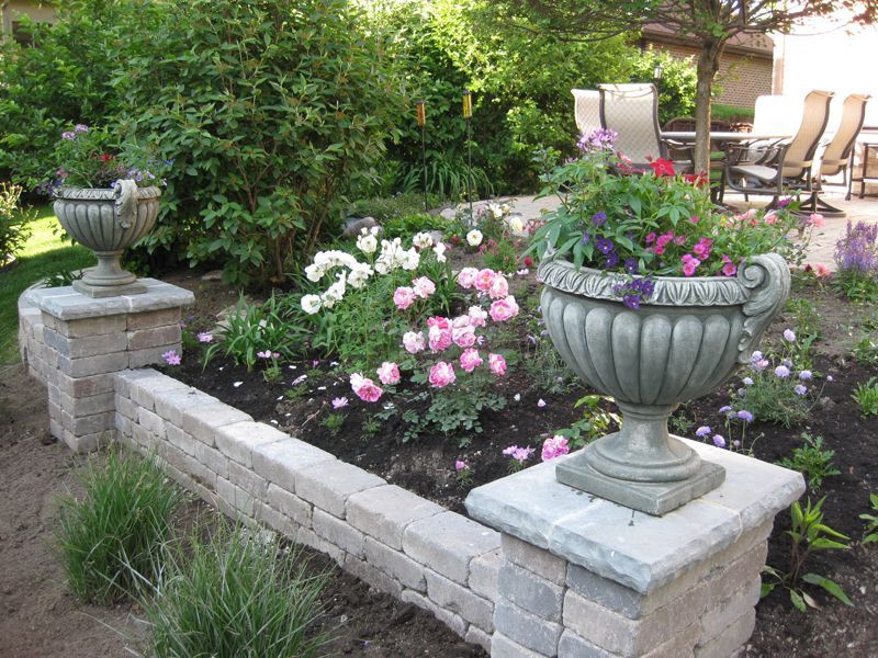 A garden bed features a stone retaining wall topped with two stone urns filled with pink and purple flowers.