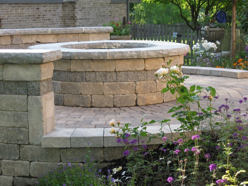 A circular stone fire pit sits on a paved patio, surrounded by a low garden wall and blooming flowers in a backyard.