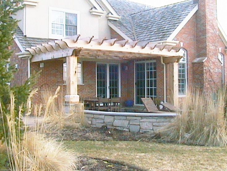 A brick house with a wooden pergola covering a stone patio, outdoor seating, and ornamental grasses in the foreground.