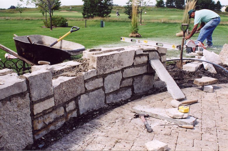 A worker constructs a stone wall on a patio overlooking a golf course, using tools and materials scattered nearby.