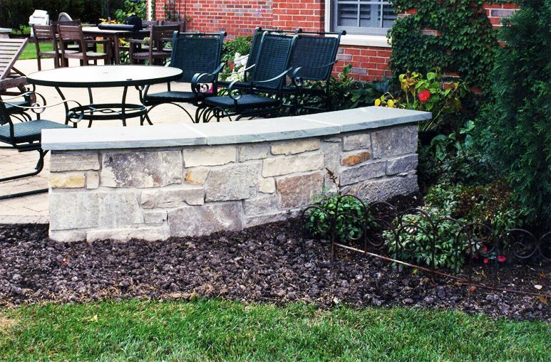 A stone wall borders a patio with a table and chairs, situated in a backyard garden with mulch and greenery.