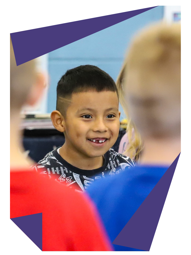 A young boy is smiling while sitting in a classroom with other children.