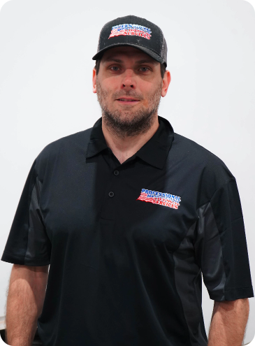 A man wearing a hat and a black shirt is standing in front of a white wall.