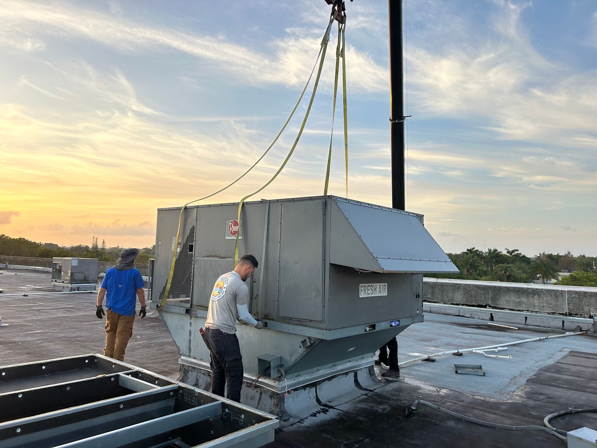 Two workers installing HVAC unit on a rooftop using a crane. Setting sun and blue sky.