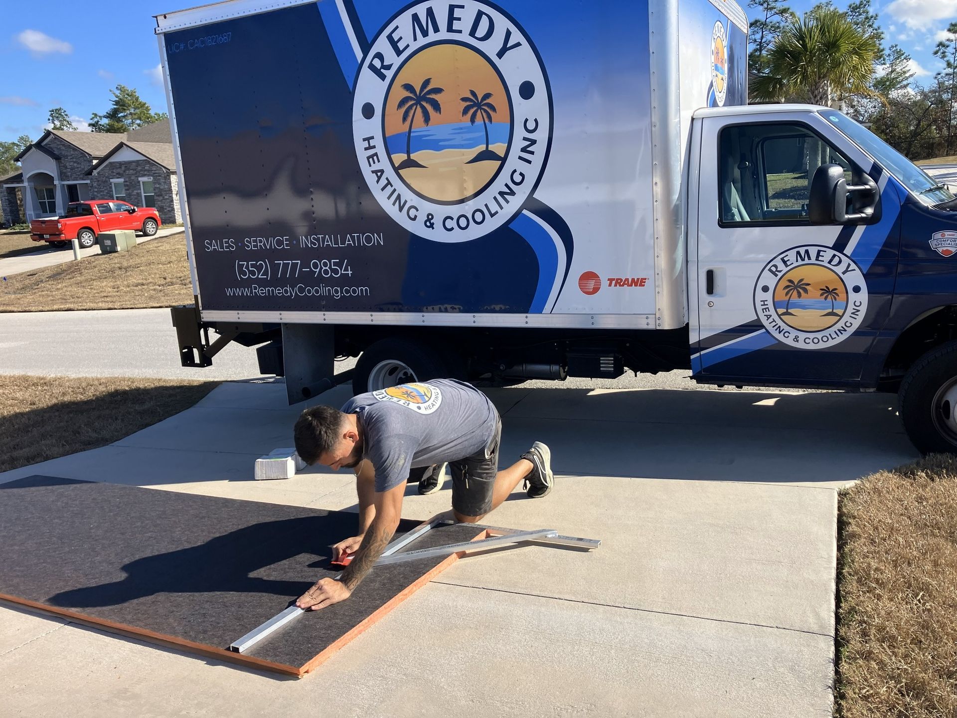 A technician kneels on a driveway, cutting material next to a Remedy Heating & Cooling truck.