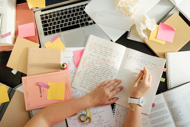 Hands writing in an open notebook on a cluttered desk with laptop, sticky notes, and other materials.