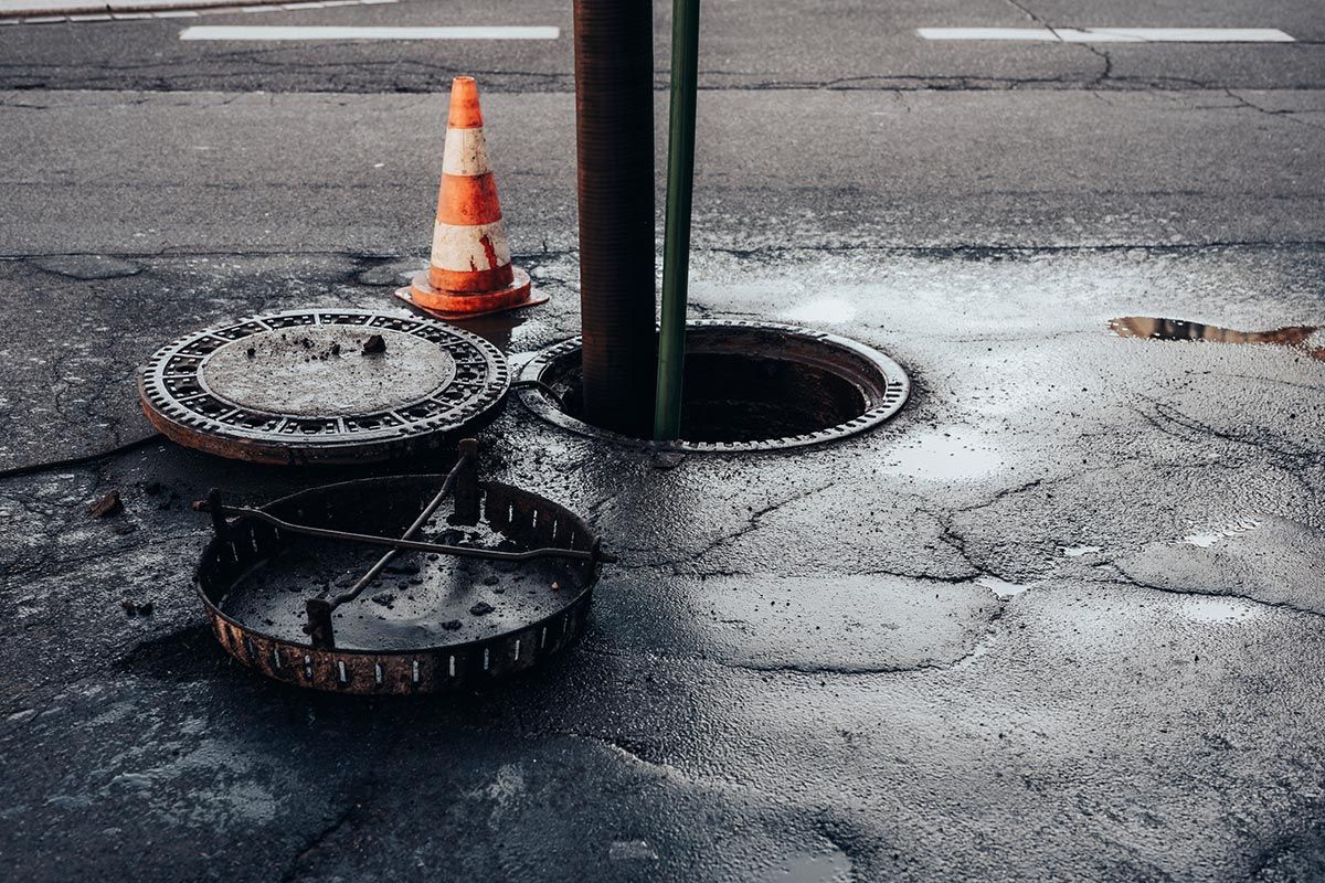 A manhole cover is open on the side of the road next to a traffic cone. A manhole cover is open on the side of the road next to a traffic cone.