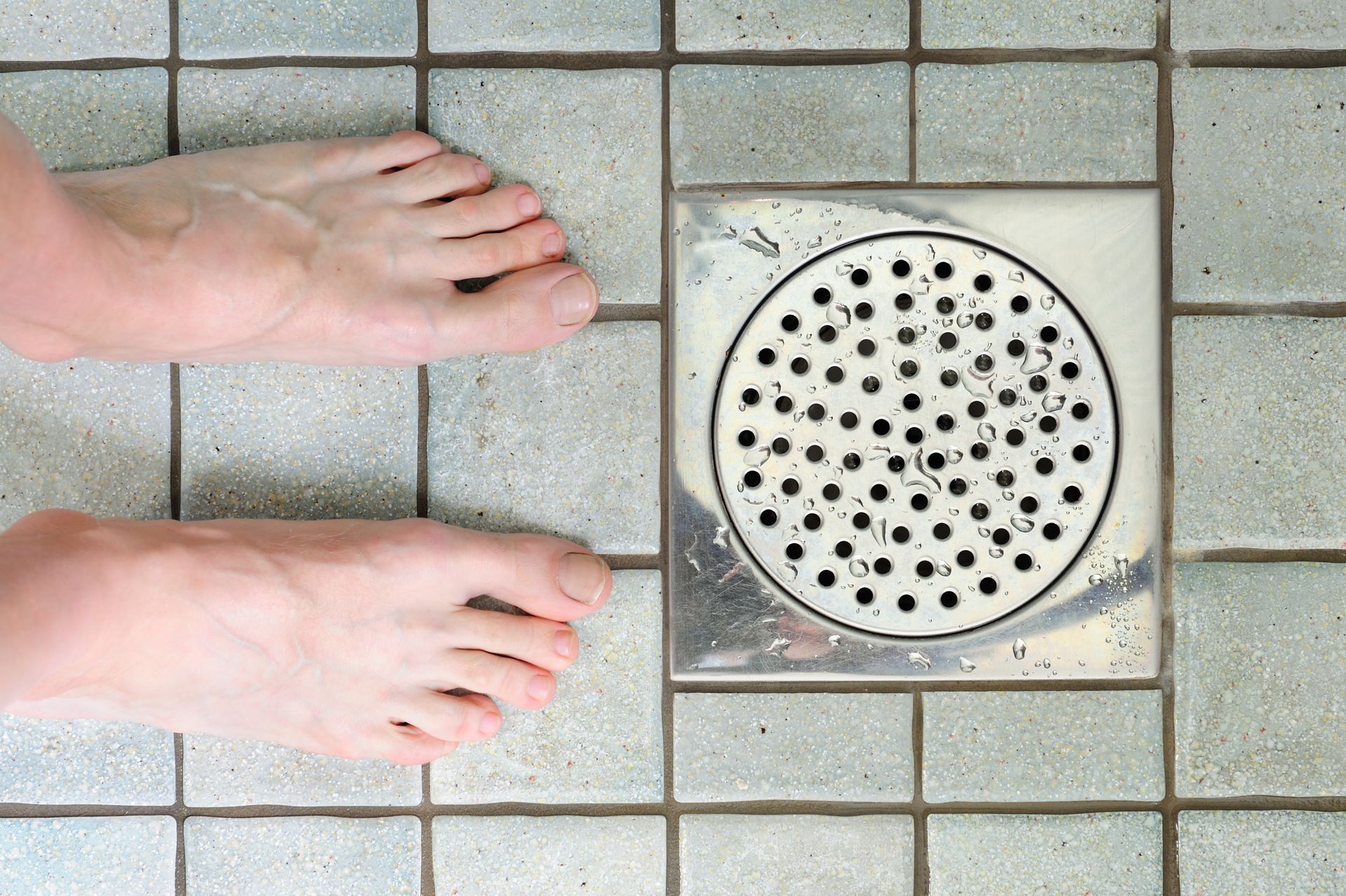 A person 's feet are standing next to a drain on a tiled floor.