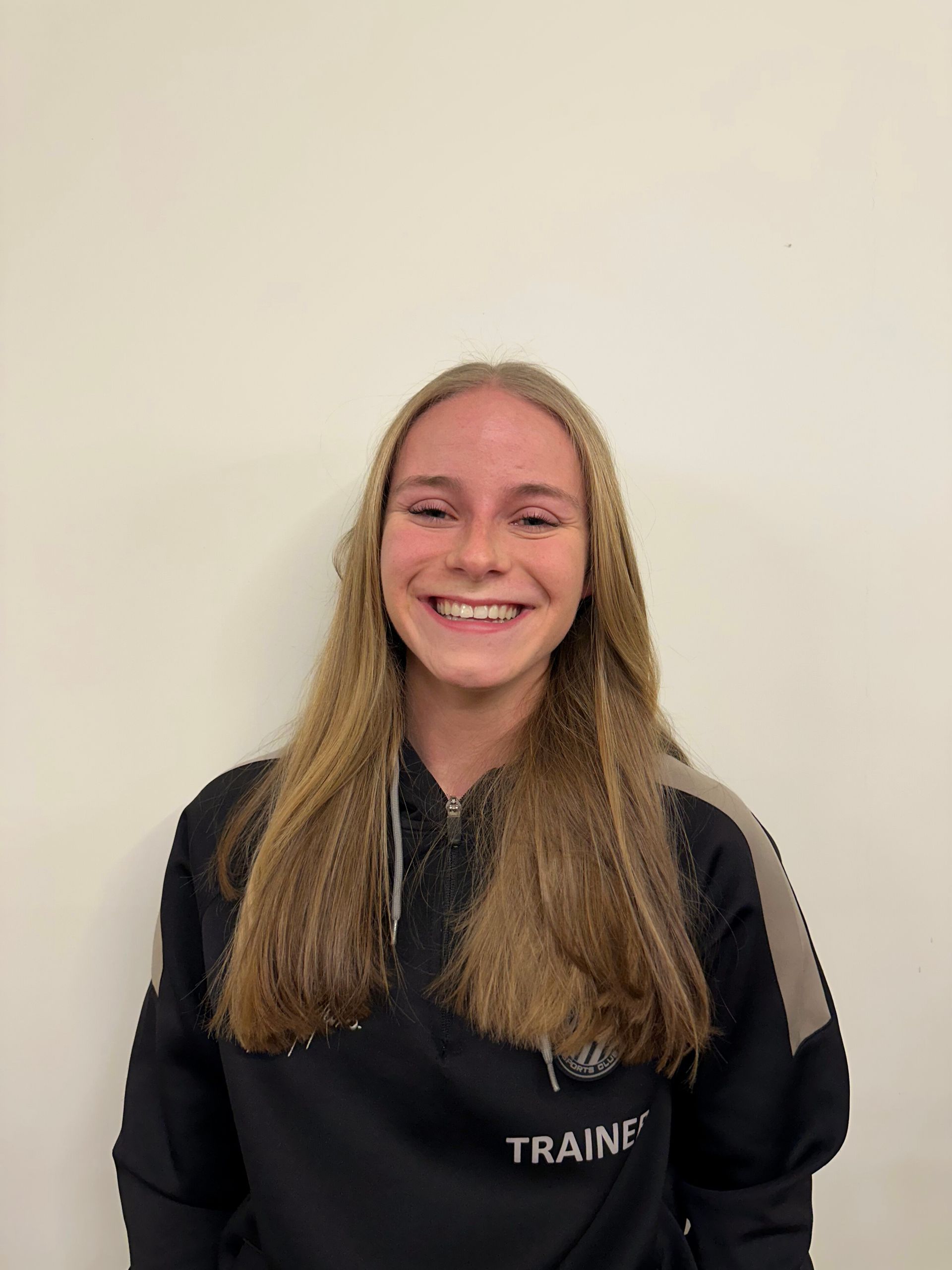 Een lachende jonge vrouw met lang blond haar en een zwart jasje staat tegen een witte muur.