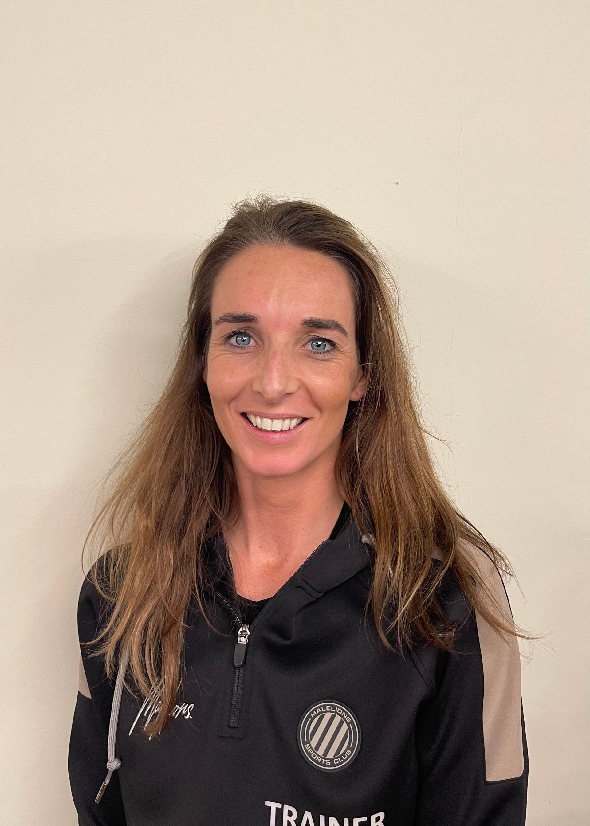 Vrouw met lang bruin haar en een zwart-beige rits, glimlachend voor een witte muur.