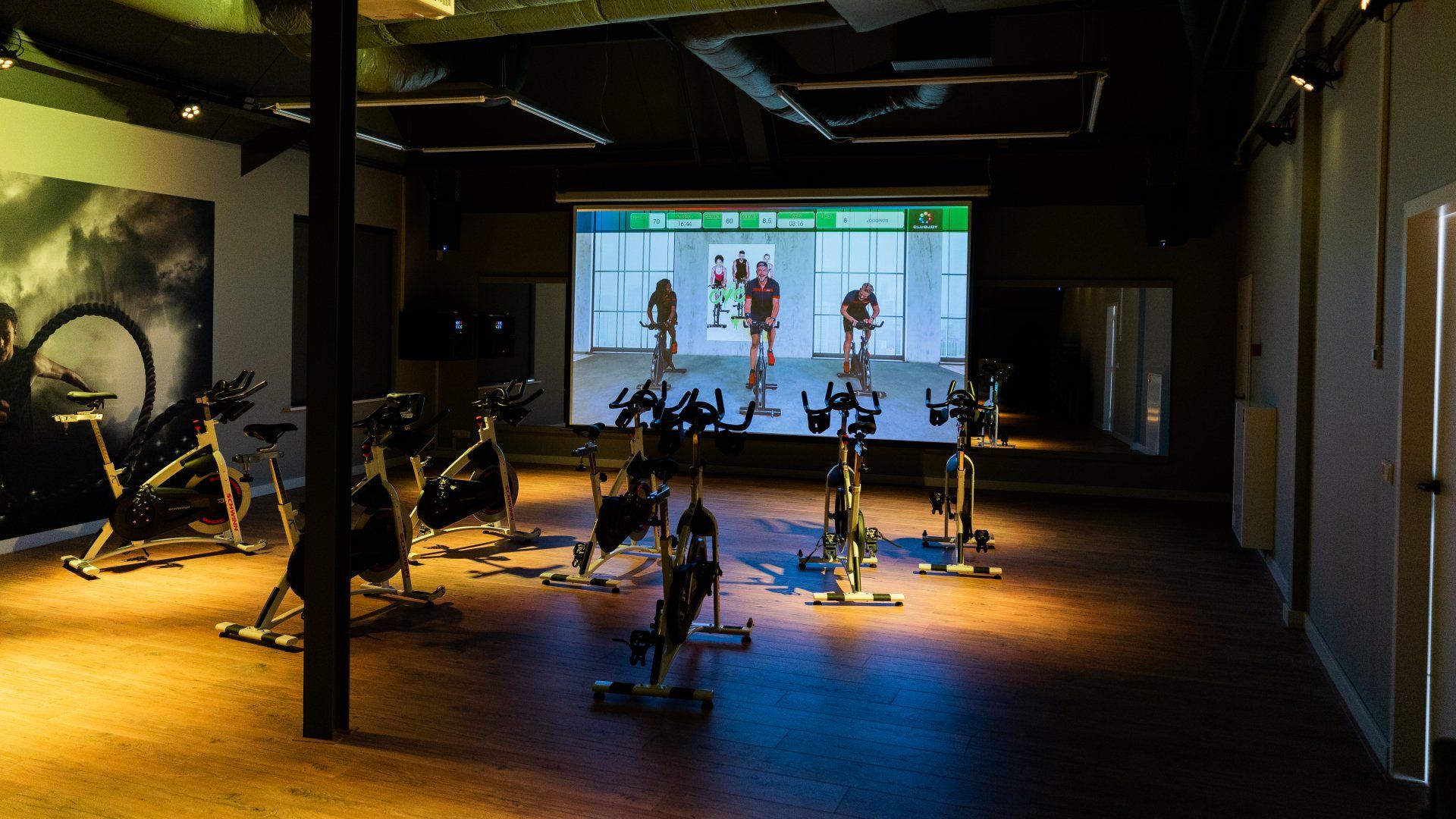 Een rij hometrainers in een sportschool met een groot scherm erachter.
