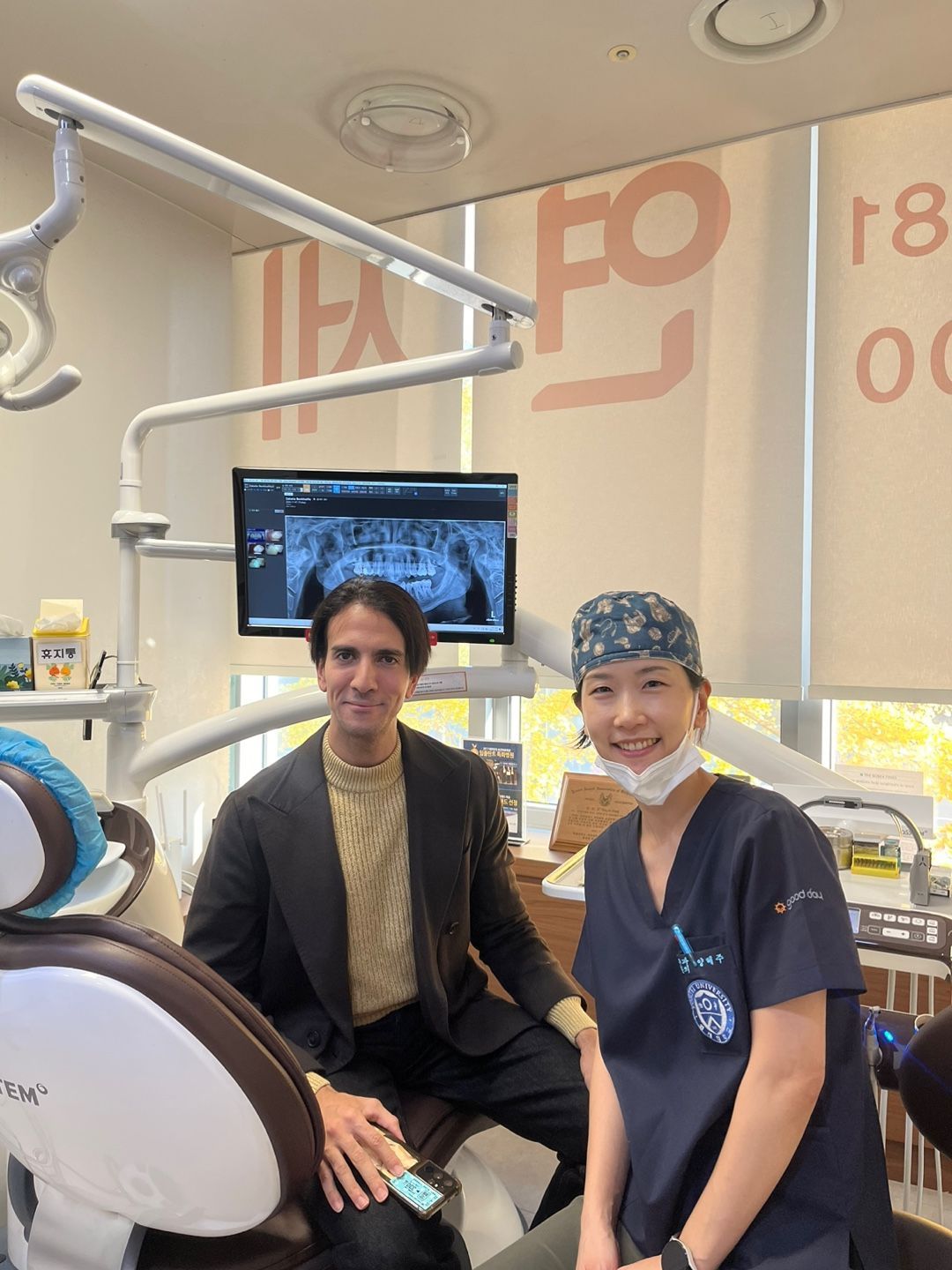 Man and dentist smiling in a dental office. The dentist wears scrubs and a patterned cap. A dental X-ray is on the monitor.