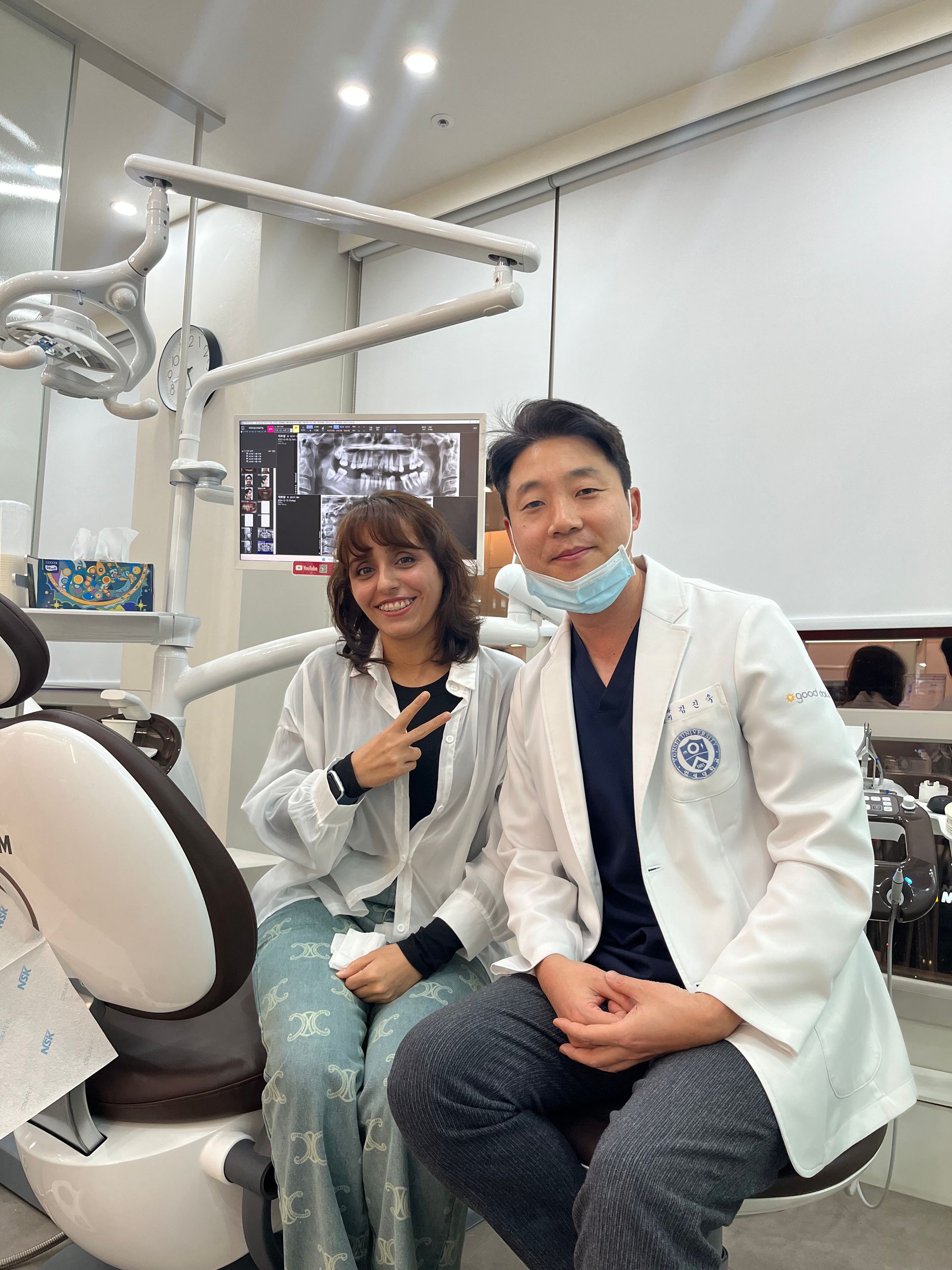 A woman and a dentist smile together in a dental office. The woman gives a peace sign. The dentist wears a mask and lab coat.