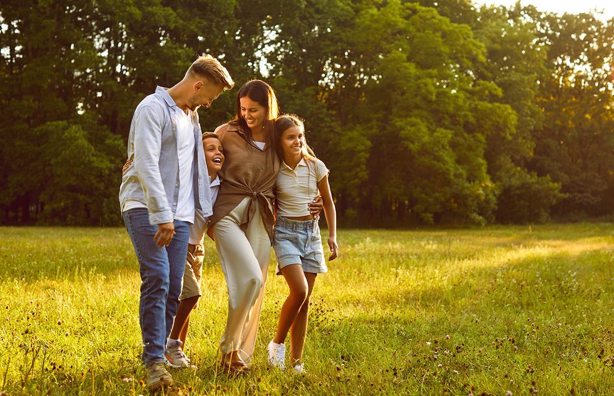 Family walking in a sunny field: mother, two children, and a teenager, green grass and trees.