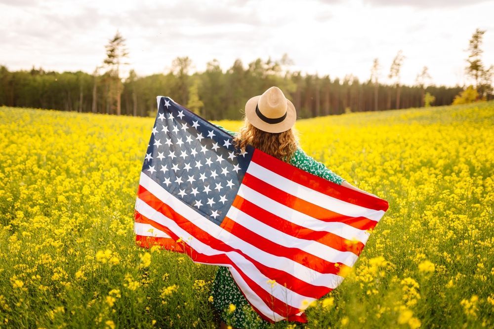 A woman is holding an american flag in a field of yellow flowers.