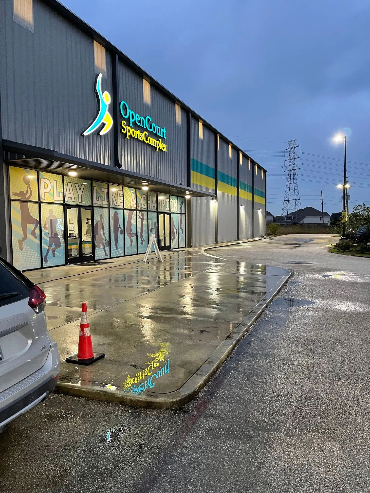 Open Sport Sports Complex exterior. Rain-slicked ground reflects building's green, yellow, and blue accents. Car parked nearby.