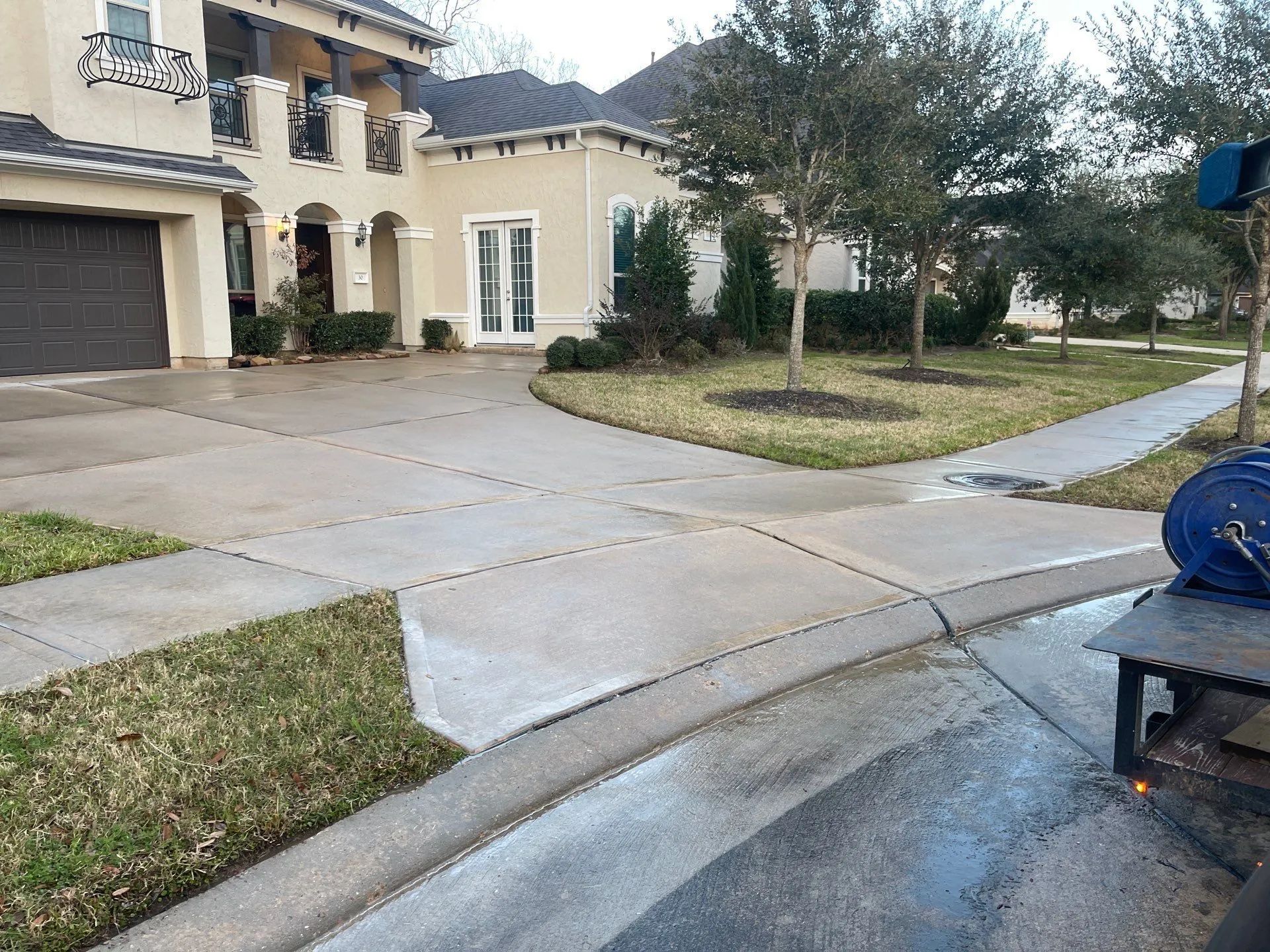 Wet concrete driveway in front of a tan two-story house, lawn and sidewalk. Pressure washer in foreground.