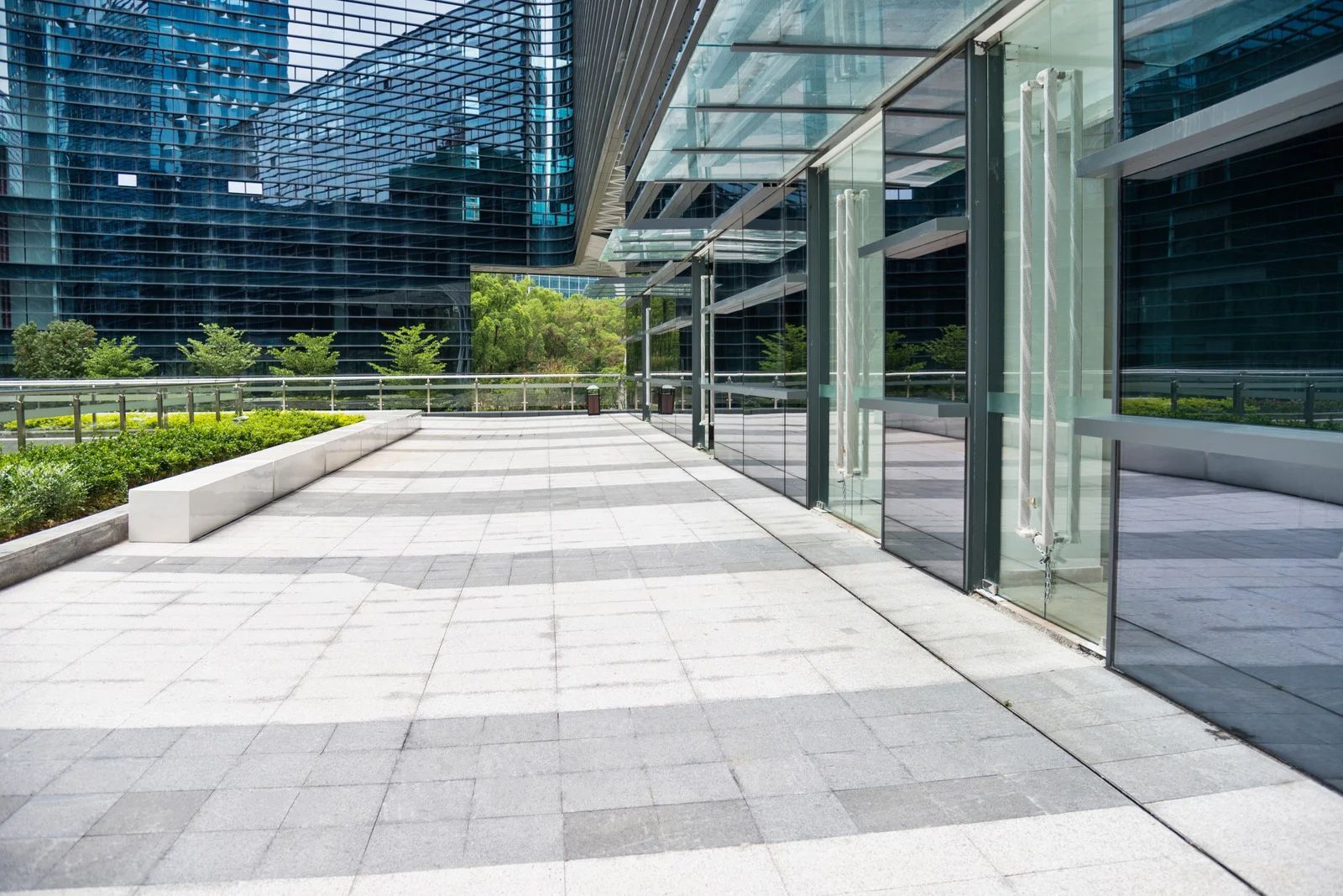 Paved walkway alongside a modern building with glass walls, greenery and tall windows visible.