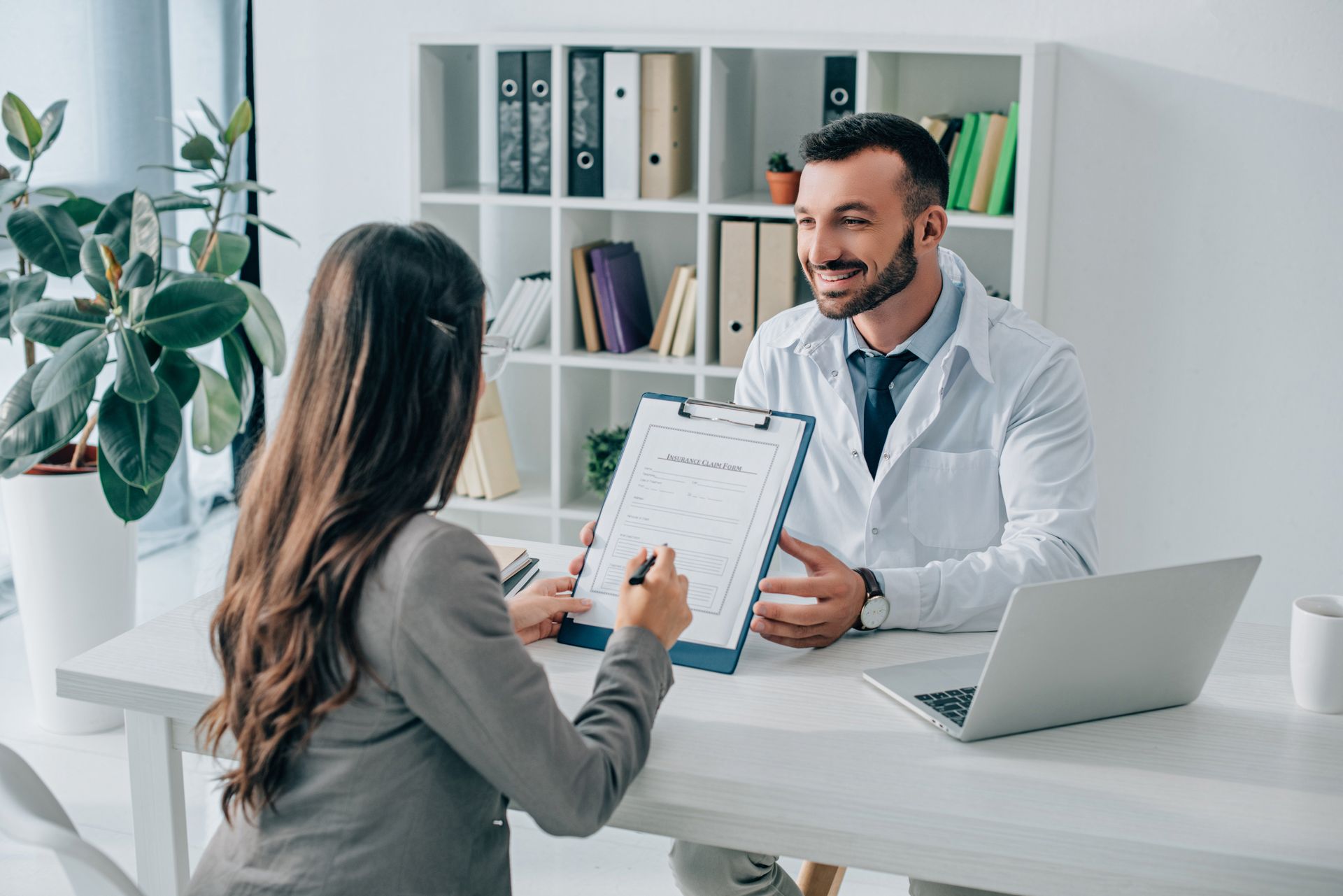 Doctor speaking with a patient across a desk in a bright clinic, reviewing a clipboard beside a laptop.