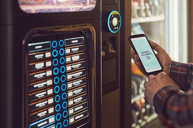 A person holds a smartphone toward a vending machine, likely using a mobile payment app to make a purchase.