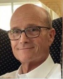 A man wearing glasses and a suit and tie is standing in front of a green curtain.