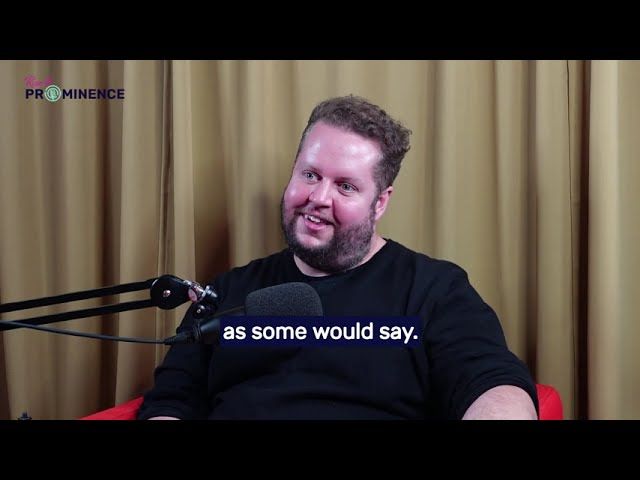 Man in black shirt smiles while speaking into a microphone. Red chair, curtains visible.