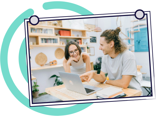 Two people at a desk, looking at a laptop and smiling. Light, airy room with books and plants.