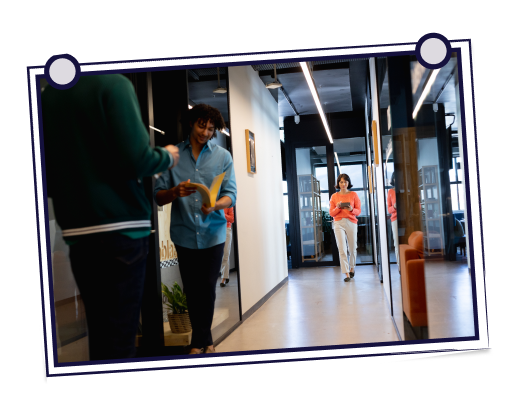 Woman waving at computer, man looks on. Both smile, working in bright room, white and yellow tones.