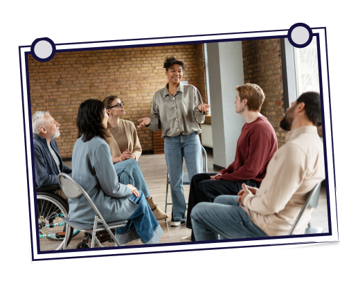 Woman at a yellow computer waves, man smiles, leaning in. Interior with plants, desk, and bed.