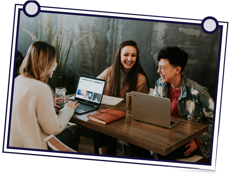 Three people at a table laughing, looking at laptops. Brown table and neutral wall background.