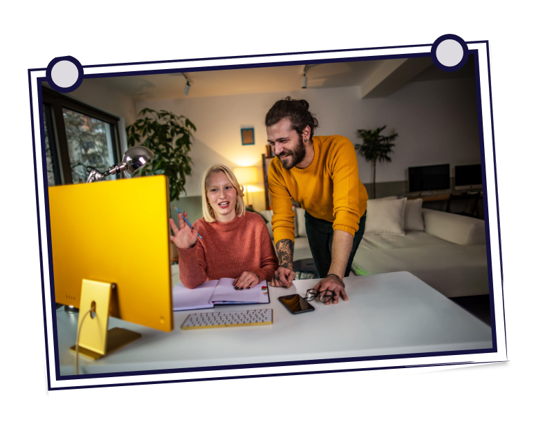 Woman waving at a computer screen, man leans over her, smiling. Desk in a living room, yellow monitor.