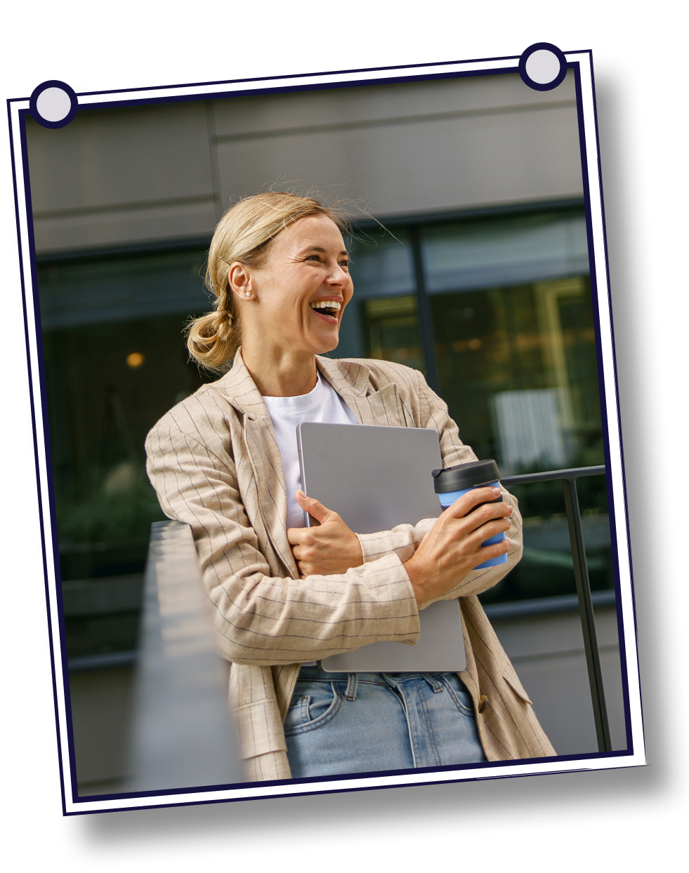 Woman laughing, holding a laptop and coffee. Wearing a beige blazer, standing outside a modern building.