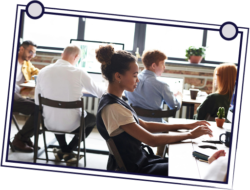 People working at desks in a bright office. Woman typing, others are looking at screens or working.