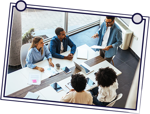 Team in a meeting at a table, discussing documents. One person stands. Bright setting with large window.