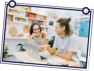 Group of people in a meeting. A man stands, gesturing while holding papers. Others seated at a table, looking at him.