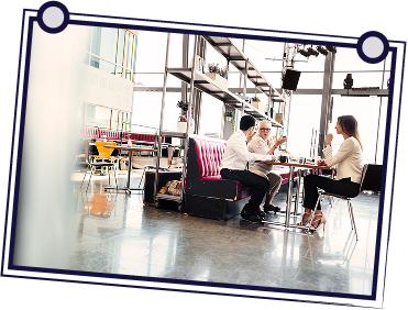 People in a meeting at a table, one person standing, gesturing while holding papers.