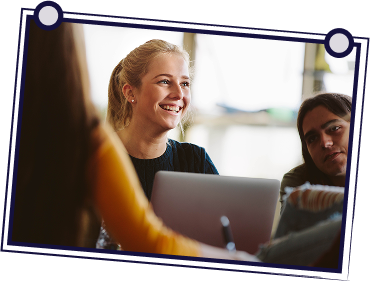 People in a meeting at a table, discussing documents. One person stands and speaks.
