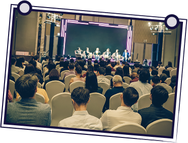 Group of people in a meeting. A man stands, gesturing while holding papers. Others seated at a table, looking at him.
