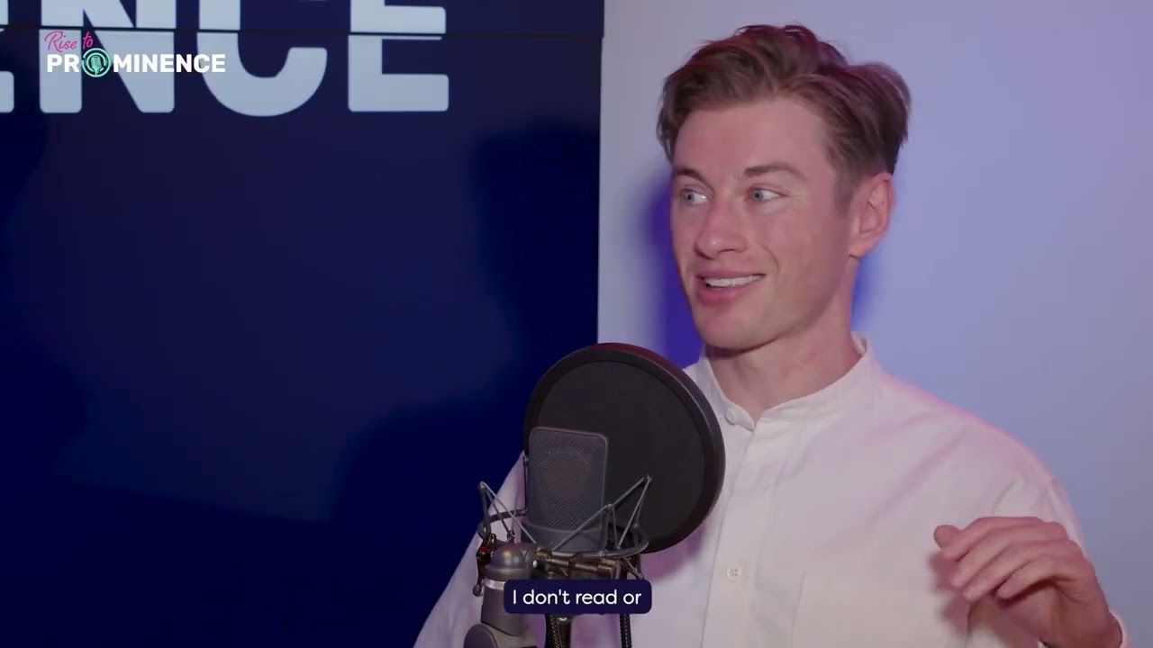 Man in white shirt speaking into microphone. Blue backdrop with "Prominence" text.