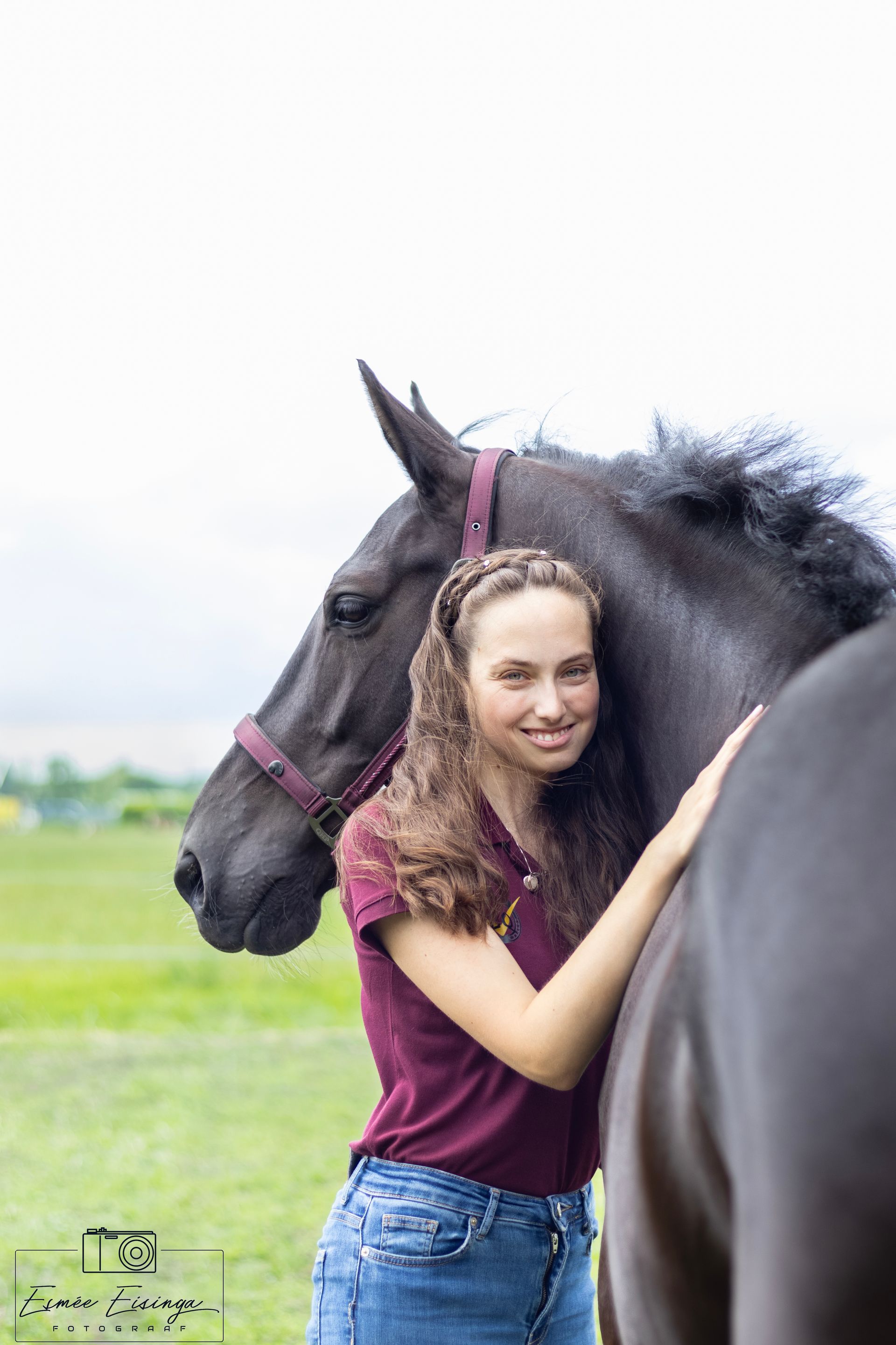 Een vrouw met lang bruin haar lacht terwijl ze een zwart paard omarmt in een grasveld.