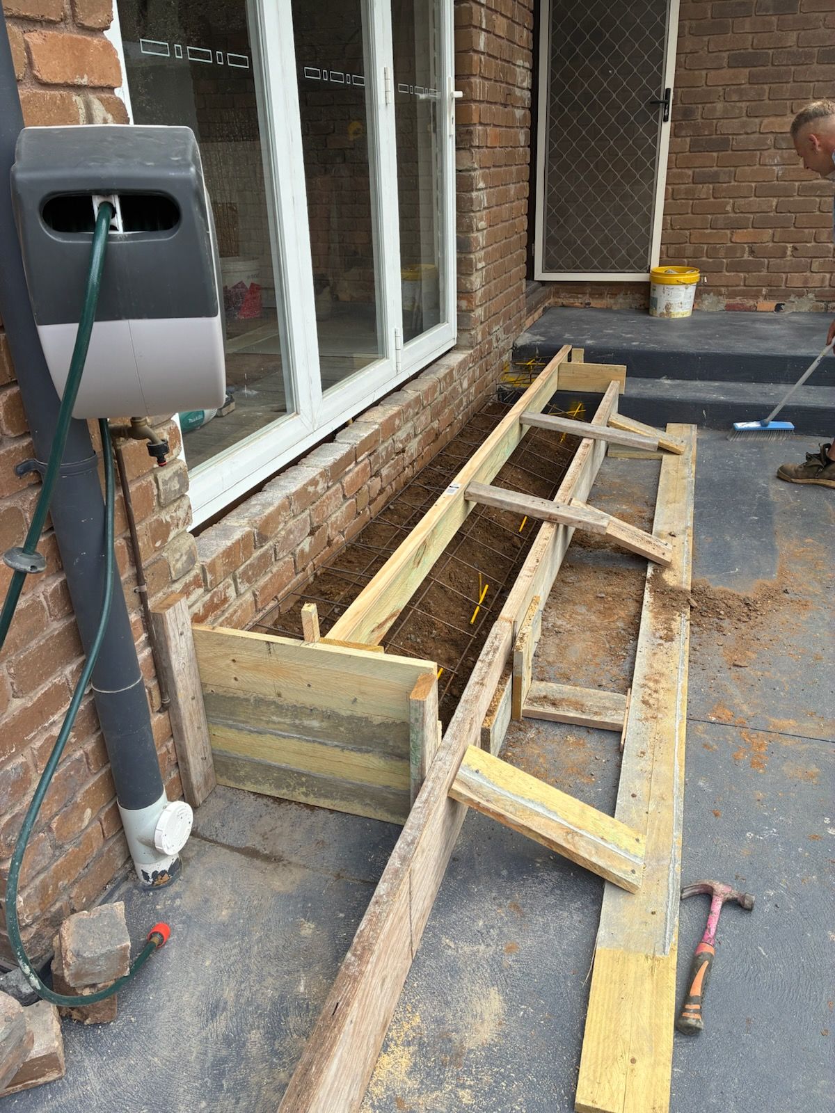 Wooden forms for concrete construction against a brick wall and window. A hose reel is on the left.