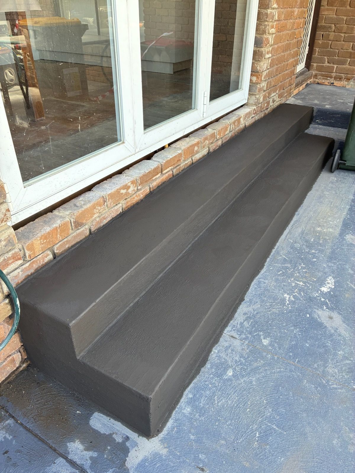 Brown, two-step outdoor stairs leading to a doorway, on a concrete patio next to a brick building.